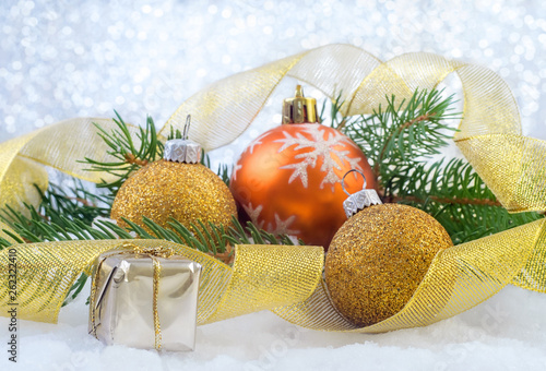 Christmas balls, small box with gifts, festive ribbon and spruce branches on the snow over a shiny background.