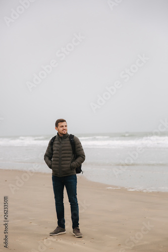 Male tourist walkin along the North Sea. Man with backpack stand on the coast in rainy weather. Beautiful sea