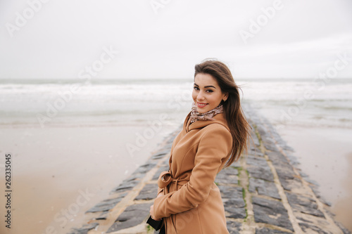 Portrait of beautiful woman walkink along the coast near the north sea. Elegant lady in brown coat with fashion make up stand in front of the sea © Aleksandr