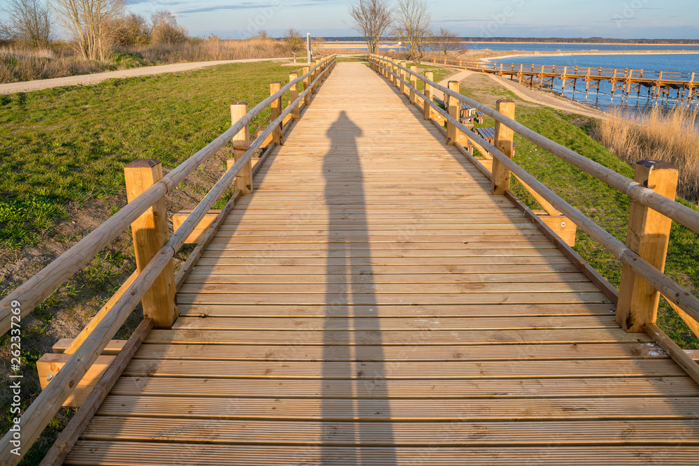 Pedestrian wooden bridge in the park. Place to rest near the lake. View of the park at sunset. Long shadow from man.
