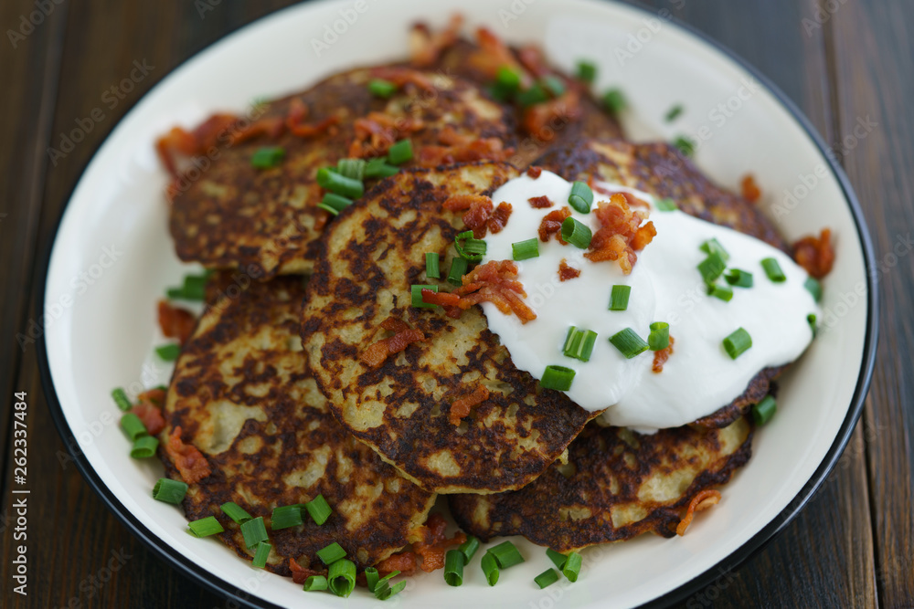 Potato pancakes draniki served with sour cream, chives and bacon on a white plate. Dark wooden table, blue linen napkin, high resolution Potato pancakes draniki served with sour cream, chives and bacon on a white plate. Dark wooden table, blue linen napkin, high resolution
