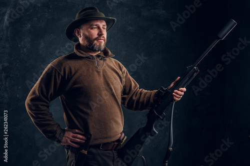 Confident hunter posing with a rifle and looking sideways. Studio photo against dark wall background