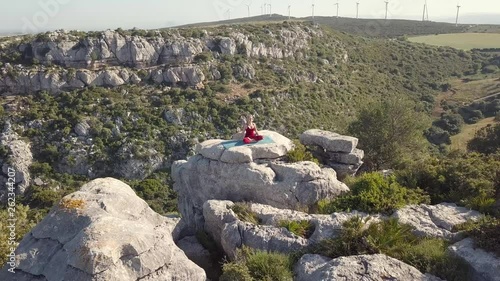 Aerial Footage of Mature Yoga Fitness Woman Performing Yoga Pose (Asana), Sitting on a Cliff at Edge of Ravine. Windmills in Distant Background. Shot Circling Left Around Model.