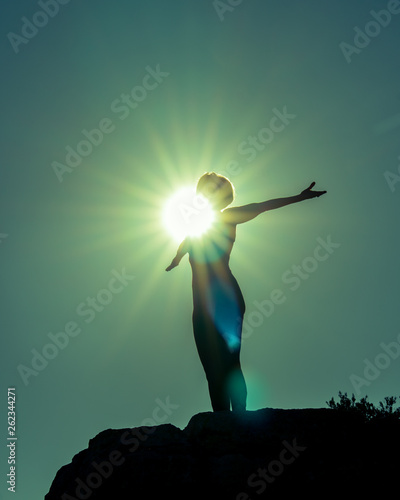 Silhouette of Yoga Fitness Woman Standing on Rock Performing Angel Pose (Asana) . She is Back Lit. Sun Flare Hitting Her Chest and Face in Center of Image. Copy Space.