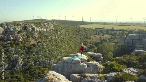 Aerial Footage of Mature Yoga Fitness Woman Performing Tree Pose (Vrikshasana ) on Cliff Edge of Ravine. Windmills in Background. Camera Moving Backwards Away from Model.