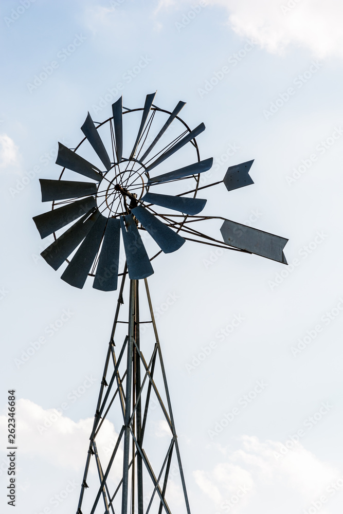 Low angle view of an old-fashioned, multi-bladed, metal wind powered water pump atop a lattice tower in backlight against a pale blue sky with white clouds. Low angle view of an old-fashioned, multi-bladed, metal wind powered water pump atop a lattice tower in backlight against a pale blue sky with white clouds.
