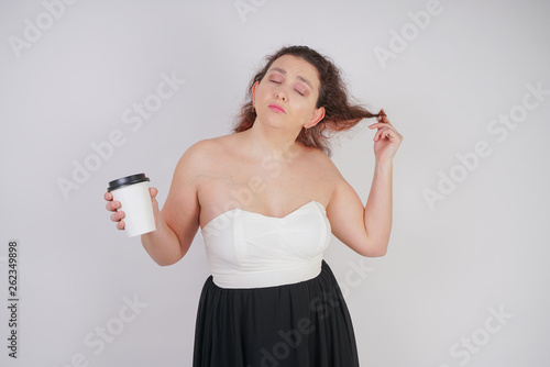 feminine woman with plus size body in a fashionable dress holding a paper Cup of coffee and posing on a white background in the Studio