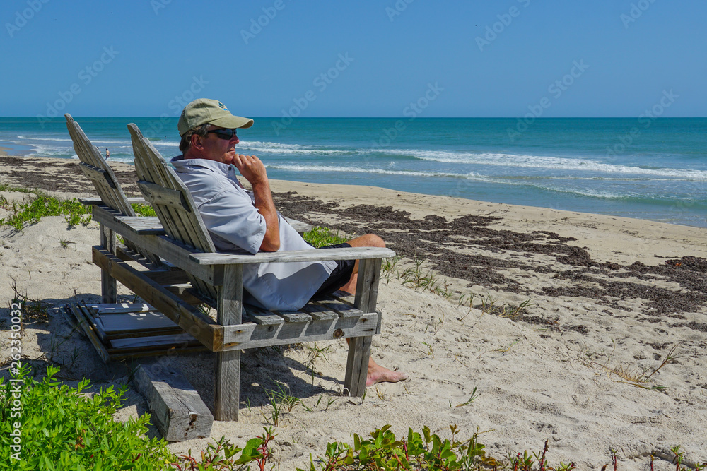 Retired man sitting on a chair overlooking the turquoise blue ocean on a bright sunny day.