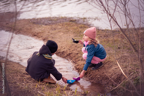 cute cheerful smiling children of 7-8 years old, curly blonde girl and boy in a coat in early spring have paper boats in the creek