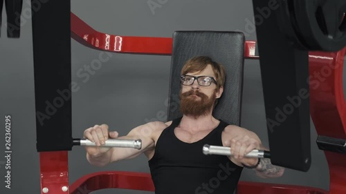 Bodybuilder in glasses doing exercise on simulator in gym. Exercise for muscles of chest. Redhead man with sports figure doing exercise on simulator in gym. Sport concept photo