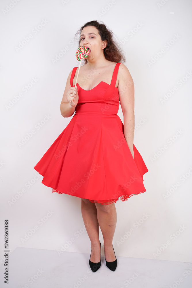 pretty chubby fashion girl wearing red pinup dress and posing with tasty colorful lollipop on white studio background pretty chubby fashion girl wearing red pinup dress and posing with tasty colorful lollipop on white studio background