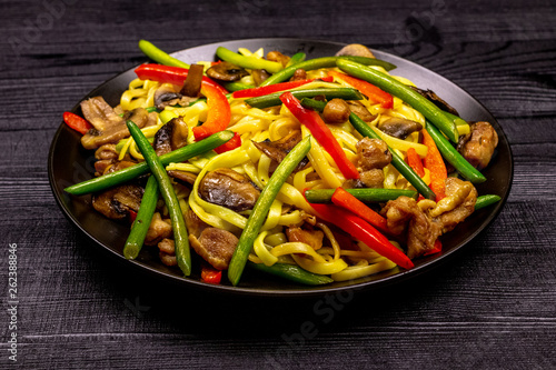Appetizing stir fried noodles with garlic sprouts, mushrooms, red pepper and chicken meat on a black rustic background of a wooden table. A popular Asian dish