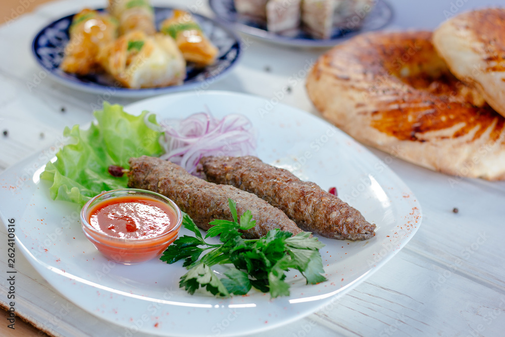 Meat kebabs on white plate with green herbs and red sauce.