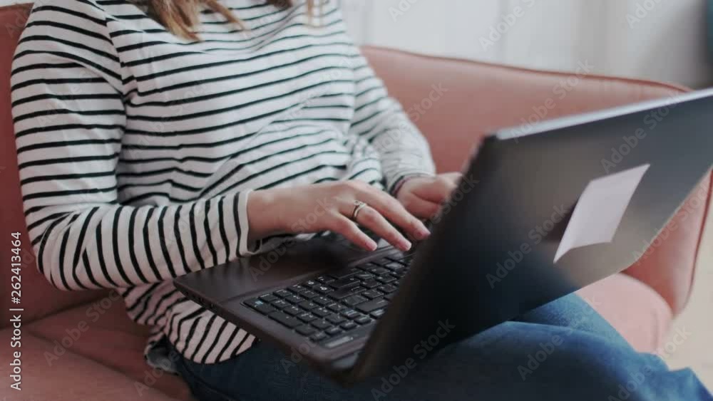 Young girl sits on sofa with laptop in daylight interior. Freelancing, working at home, checking posts, write comments in social media. Female hands type on laptop keyboard touching touchpad Young girl sits on sofa with laptop in daylight interior. Freelancing, working at home, checking posts, write comments in social media. Female hands type on laptop keyboard touching touchpad
