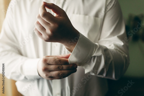 Stylish groom preparing for wedding ceremony, handsome man buttoning white shirt in hotel room, hands closeup, tailor concept