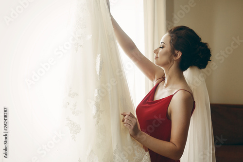 Beautiful bride in red robe with veil looking at elegant white lace wedding dress on hanger near a window, morning wedding preparation in hotel