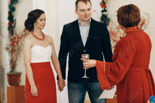 Mother in law pronouncing toast to happy newlywed couple, woman with champagne glass drinking to bride and groom at wedding ceremony in registry office photo