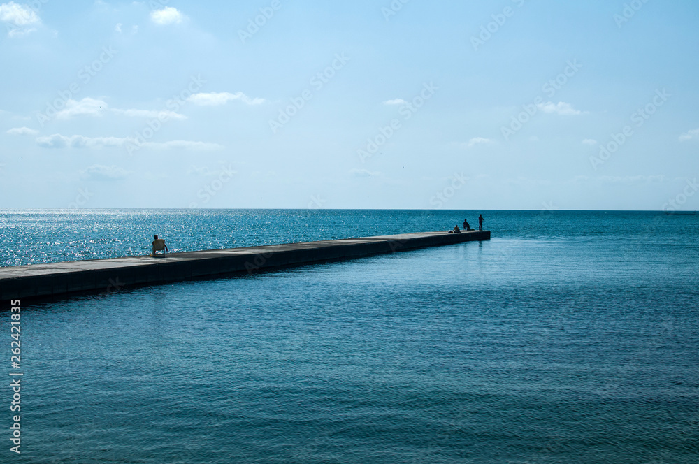 Stone quay in sea bay of calm blue sea waters and three human silhouettes in clear sunny summer day