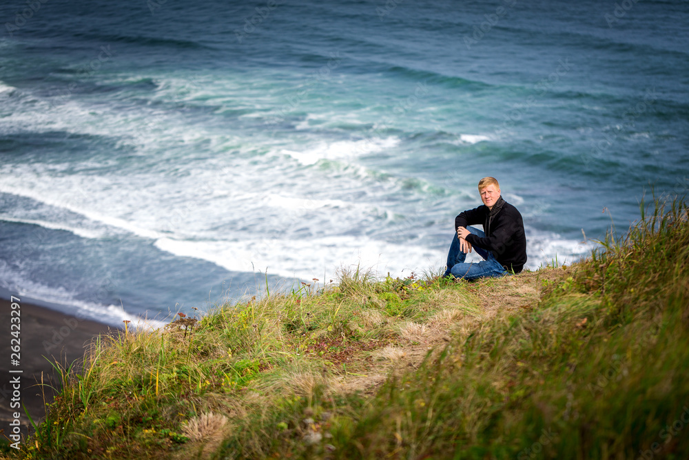 Redhead man in a black sweater sits on the side of a hill near the Khalaktyrsky black beach on the Kamchatka Peninsula, washed by the Pacific Ocean. Travel, hiking and adventure concept.