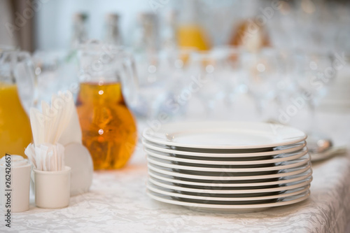 A stack of white plates on the table with a carafe of juice plastic cups