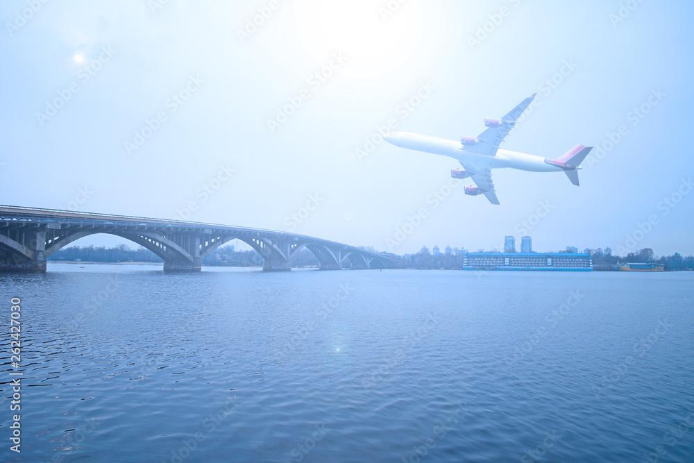 Naklejka premium The plane over the river and the bridge in the city. Cloudy day.