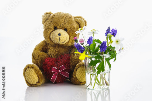 Ein Teddybär mit Herz neben einer Vase mit Blumen. 