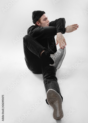 Freestyle dancer dressed in black jeans, sweatshirt, hat and gray sneakers is dancing sitting on the floor in the studio on the white background