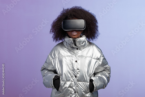 Modern curly brown-haired girl dressed in a silver-colored jacket uses the virtual reality glasses poses  in the studio on neon background photo