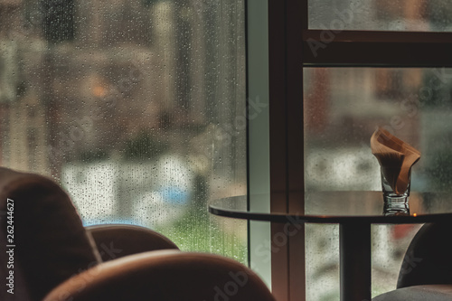 Design interior with coffee table and armchair near the window. Raindrops on the window glass. Blurred cityscape on background. Overcast rainy weather. Selective focus on window. 