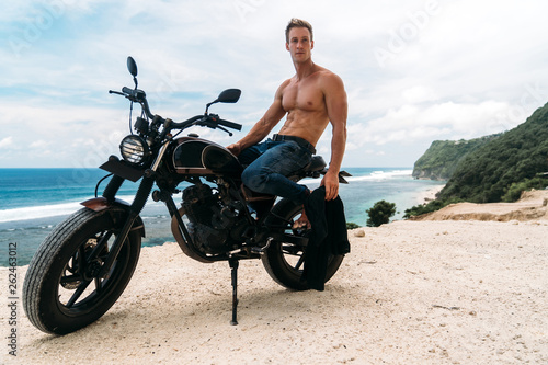 Sexy athletic man with perfect naked body sitting on motorbike, ocean waves and beautiful mountains on background