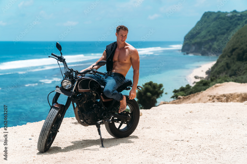 Obraz premium Sexy athletic man with perfect naked body sitting on motorbike, ocean waves and beautiful mountains on background