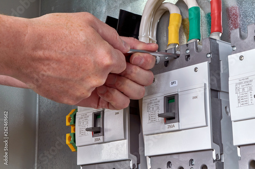 A person or worker screws an electric circuit breaker with a key to the circuit Board in the electrical Cabinet. Three powerful switches are fixed in a row. Switches are connected to cables or wires.