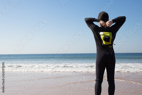 Fotografie Back view of young man putting on wetsuit on sunny beach