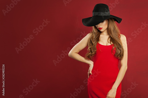 Beautiful woman fashion model wearing red dress and black hat standing against red wall background © artmim