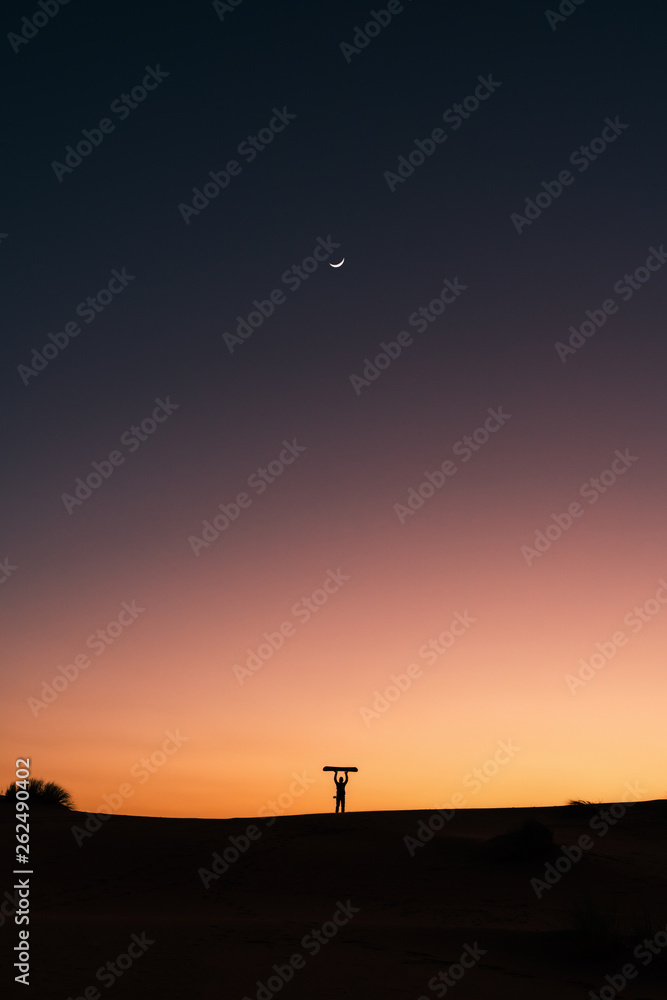Silhouette of a man with snowboard in the Sahara desert, sunset and moon