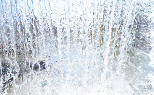 Transparent blue white water pours from above. View through the water wall of the waterfall for the background.