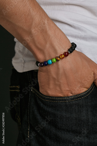 Cropped closeup half-turn shot of man's hand with multicolor stony bracelet with semiprecious stones. The guy in jeans and white T-shirt is putting his hand into pocket, standing on dark background. photo