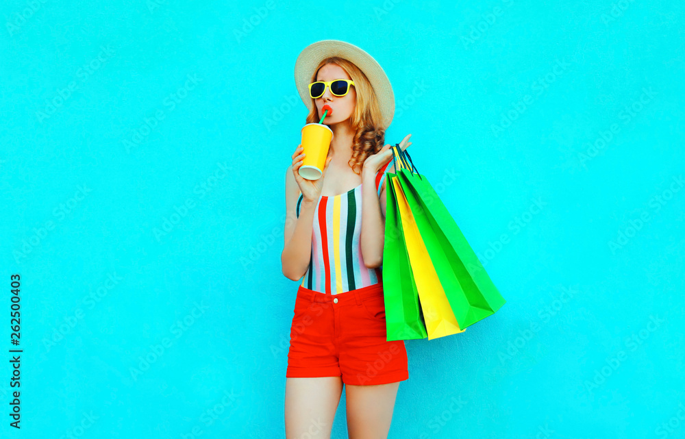 Stylish woman drinking juice holding shopping bags in colorful t-shirt, summer straw hat, sunglasses, red shorts on blue background Stylish woman drinking juice holding shopping bags in colorful t-shirt, summer straw hat, sunglasses, red shorts on blue background