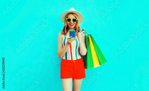 happy smiling young woman holding smartphone with shopping bags in colorful t-shirt, summer straw hat, sunglasses, red shorts on blue background