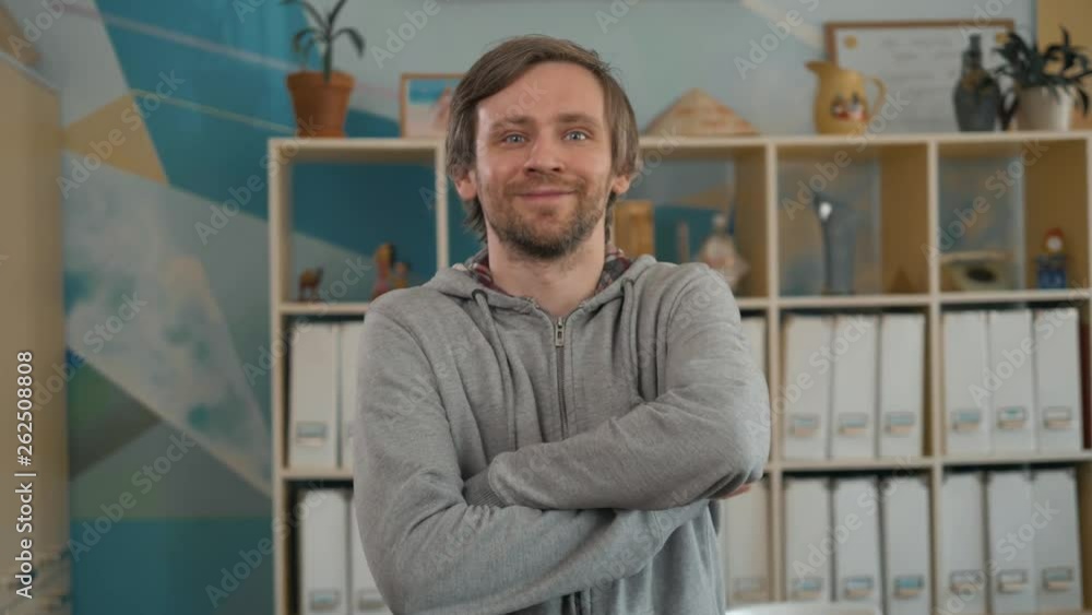 Portrait of handsome young man standing with his arms crossed in creative office. Confident young male creative professional looking at camera and smiling coworking colorful place Portrait of handsome young man standing with his arms crossed in creative office. Confident young male creative professional looking at camera and smiling coworking colorful place