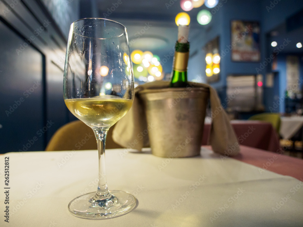 Wide closeup of a glass of Alsatian white wine as an aperitif at a romantic restaurant dinner. Shallow focus, bucket and chilled bottle on table in background. Colmar, France. Travel and cuisine. Wide closeup of a glass of Alsatian white wine as an aperitif at a romantic restaurant dinner. Shallow focus, bucket and chilled bottle on table in background. Colmar, France. Travel and cuisine.