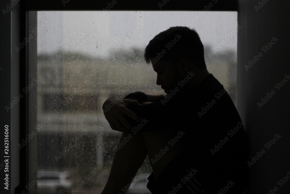 silhouette of a man sitting in the window with rain in mood of sadness and depression