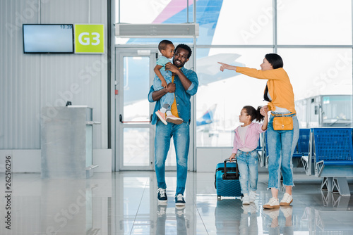 smiling african american family walking with baggage along waiting hall in airport while mother pointing with finger at son