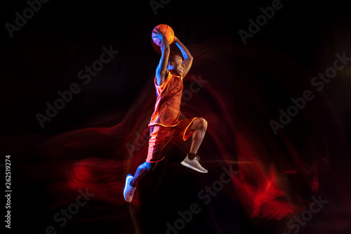 Fototapeta Naklejka Na Ścianę i Meble -  One step higher. African-american young basketball player of red team in action and neon lights over dark studio background. Concept of sport, movement, energy and dynamic, healthy lifestyle.