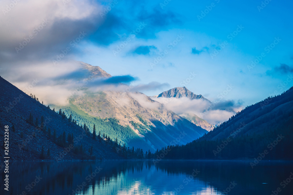 Amazing blue silhouettes of mountains under blue cloudy sky. Beautiful ripples on water of mountain lake. Low clouds before mountain ridge. Wonderful highland landscape. Picturesque mountainscape.