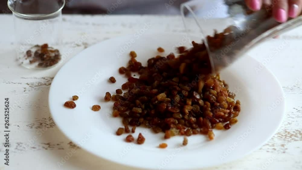 A young housewife pours raisins on a white plate a glass cup on a light wooden table in the kitchen. Women's hands. Close-up. A young housewife pours raisins on a white plate a glass cup on a light wooden table in the kitchen. Women's hands. Close-up.