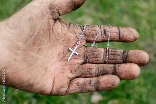Silver cross in dirty hand of homeless elderly man on green background. Salvation of man, help, faith in Jesus, Holy Easter.
