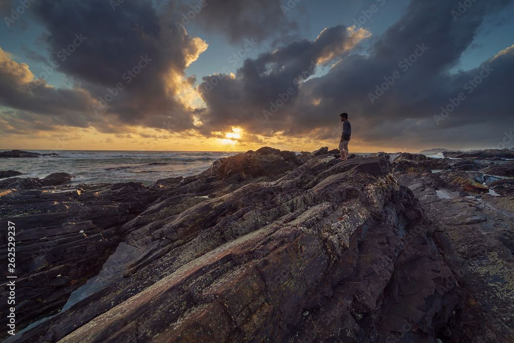 A man standing on rocks at beach side with stunning and dramatic clouds sunrise scenery at Pantai Tanjung Jara, Dungun.