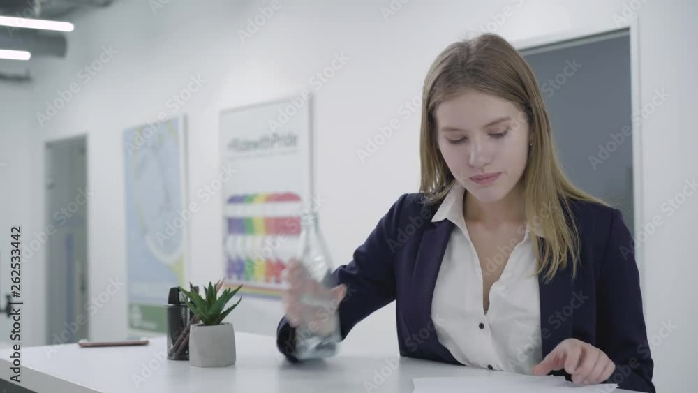 Concerned thoughtful young lady in formal clothes working with papers in the office standing at the counter. Woman with long hair takes water bottle and drinks. Working process. Workplace. Office Concerned thoughtful young lady in formal clothes working with papers in the office standing at the counter. Woman with long hair takes water bottle and drinks. Working process. Workplace. Office