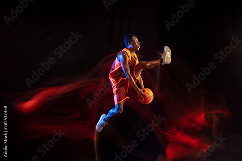 Higher and stronger. African-american young basketball player of red team in action and neon lights over dark studio background. Concept of sport, movement, energy and dynamic, healthy lifestyle.